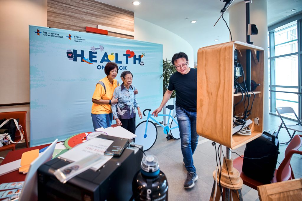 Guests posing for a photo booth in front of a Health on Track event backdrop, with the booth operator at a wooden camera housing