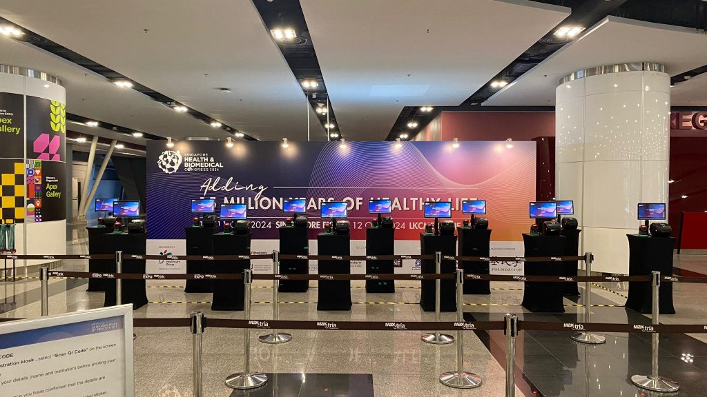 Row of self-service registration kiosks with screens and badge printers in front of a medical congress backdrop at Singapore EXPO